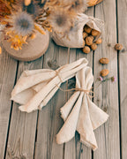 Two folded cream linen napkins tied with twine are arranged on a rustic wooden table, alongside a small fabric bag of walnuts, dried flowers in a ceramic pot, and a loaf of bread.