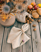 A styled flat-lay of natural beige linen fabric items on a weathered gray wooden surface, including a gathered cloth napkin tied with twine and a matching fabric bag spilling walnuts, alongside dried flowers, bread, and apples.