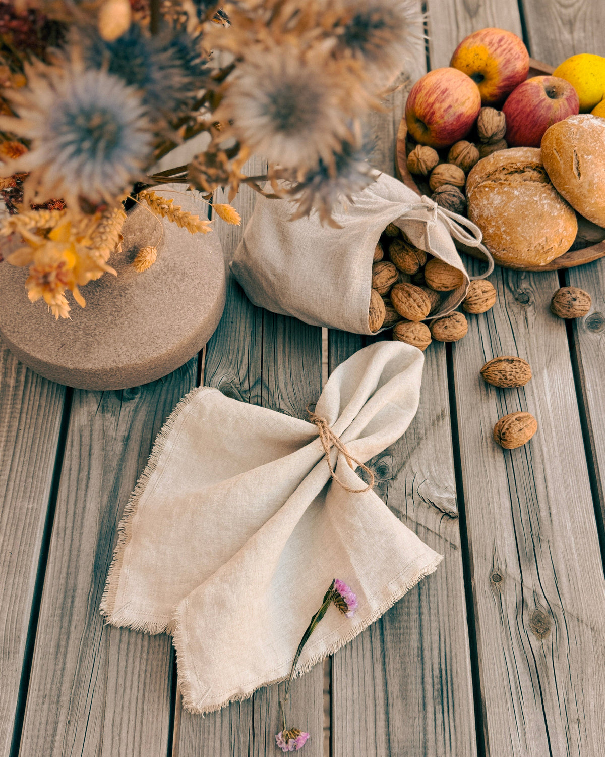 A styled flat-lay of natural beige linen fabric items on a weathered gray wooden surface, including a gathered cloth napkin tied with twine and a matching fabric bag spilling walnuts, alongside dried flowers, bread, and apples.