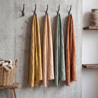Four lightweight linen scarves in mustard yellow, blush pink, sage green, and terracotta orange hang from dark metal hooks on a textured concrete wall, with a wicker basket and wooden shelf visible in a styled interior setting.