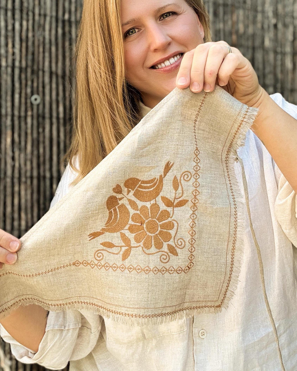 A woman holding up a square beige linen napkin with brown embroidered birds and a flower, set against a rustic wooden background.