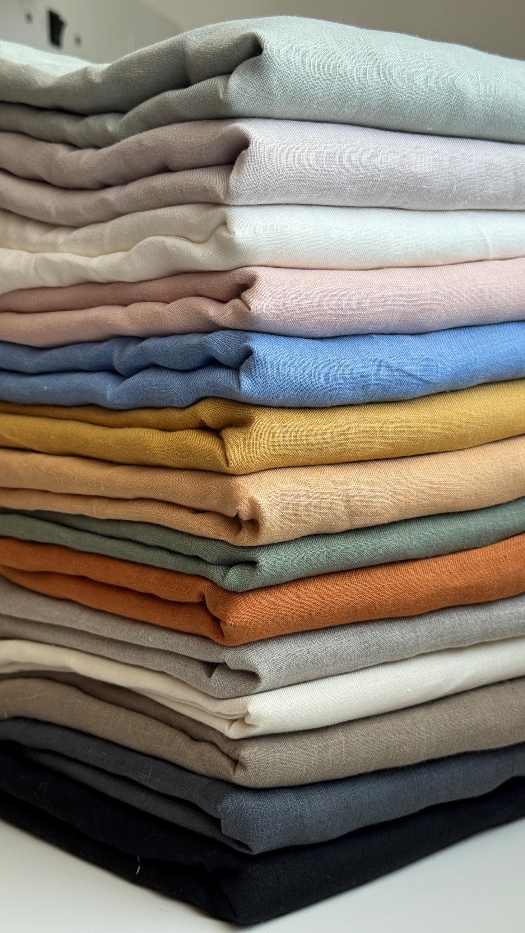 A stacked collection of folded linen fabrics in a variety of muted colors, including sage green, dusty pink, light blue, mustard yellow, terracotta orange, and charcoal gray.