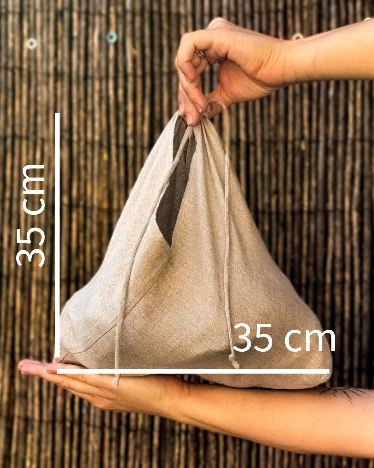 A beige linen drawstring bag being held by two hands against a dark bamboo screen, with white dimension lines and text indicating a 35 cm height and 35 cm width.