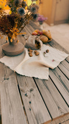 A rustic styled tabletop scene with a beige linen cloth spread across weathered wooden planks, holding a small sack of acorns, scattered walnuts, and fruit, alongside a ceramic vase of dried flowers.