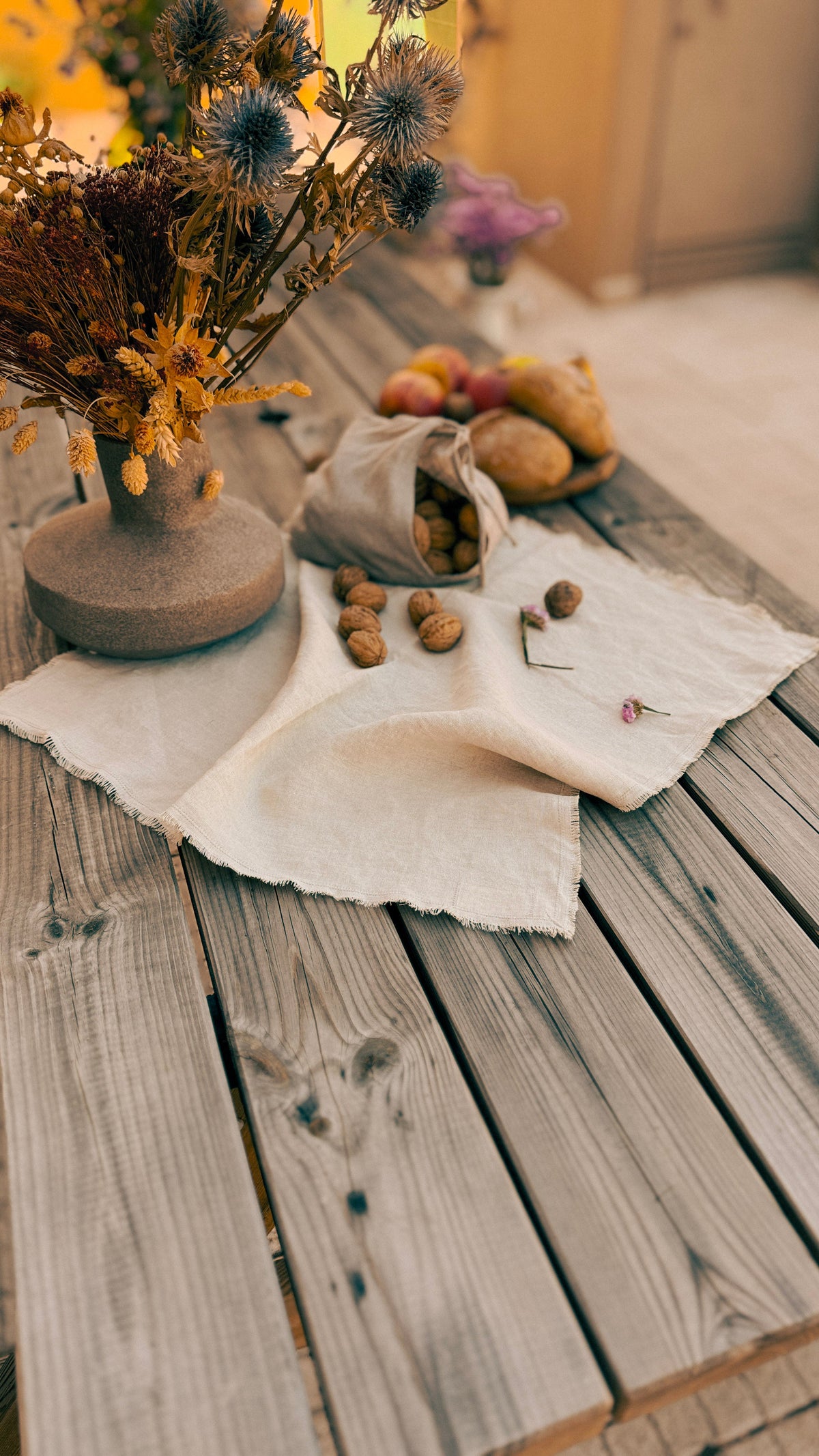 A rustic styled tabletop scene with a beige linen cloth spread across weathered wooden planks, holding a small sack of acorns, scattered walnuts, and fruit, alongside a ceramic vase of dried flowers.