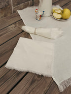 A set of off-white woven table linens with fringed edges, including a flat square napkin and a rolled rectangular cloth tied with a string, displayed on a dark wooden table alongside a white pitcher, a small rooster figurine, and sliced yellow fruit.