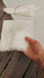 A hand holding a folded white fringed fabric napkin over a dark wooden table, with more folded napkins in the background.