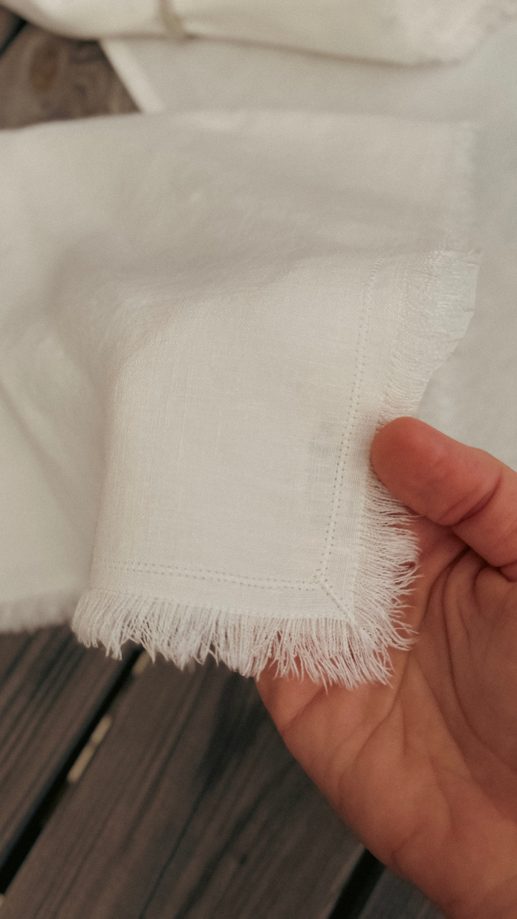 A hand holding a folded white fringed fabric napkin over a dark wooden table, with more folded napkins in the background.