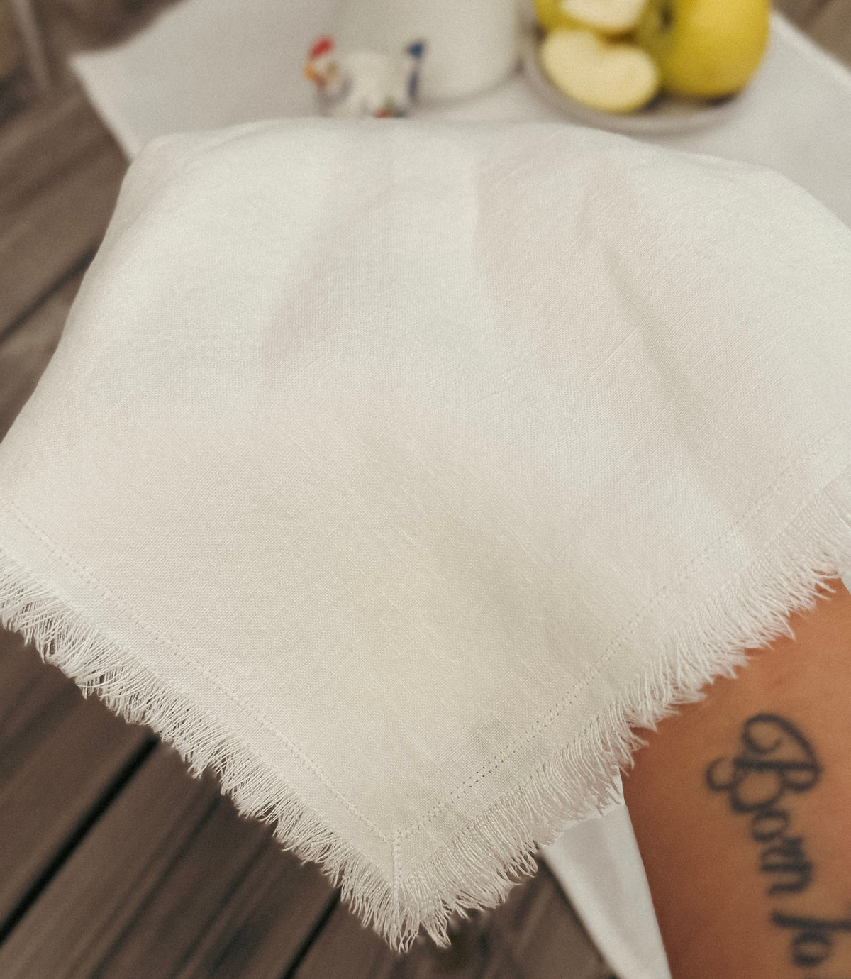 A hand holding a folded white fringed fabric napkin over a dark wooden table, with more folded napkins in the background.