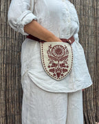 A person wearing a white linen outfit carries a small shield-shaped cream fabric pouch with a brown embroidered floral design and a brown strap, standing against a bamboo screen.