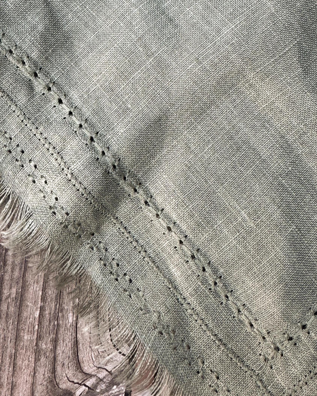 Close-up of a woven textile with a textured sage green surface, decorative perforated stitching, and fringed edges, laid over a rustic wooden surface.