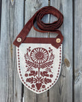 A rustic shield-shaped fabric pouch with a brown strap, featuring a brown folk-art floral and bird pattern on a cream linen background, laid flat on a gray wooden plank surface.