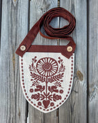 A rustic shield-shaped fabric pouch with a brown strap, featuring a brown folk-art floral and bird pattern on a cream linen background, laid flat on a gray wooden plank surface.