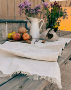 A rustic outdoor table setting featuring a light beige linen tablecloth with fringed edges, styled with fresh fruit, a white ceramic pitcher filled with purple wildflowers, and a small fabric bag.