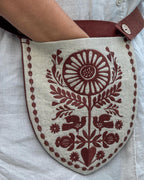 A close-up of a shield-shaped linen pouch with dark red embroidered floral and bird motifs, worn over a white shirt.