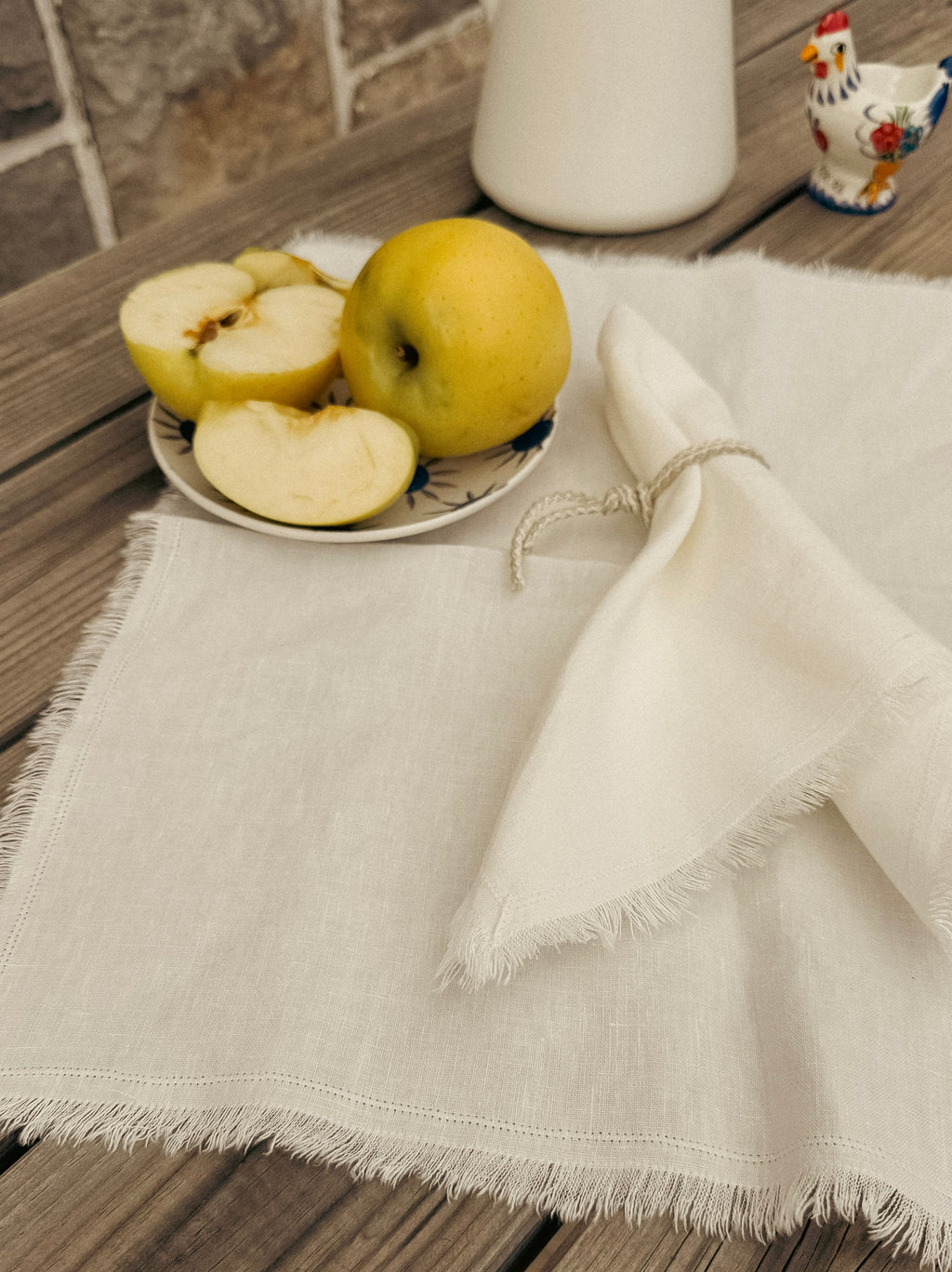 An off-white linen table runner with frayed edges draped on a rustic wooden table, styled with a matching napkin tied with twine and a small plate of yellow apples.
