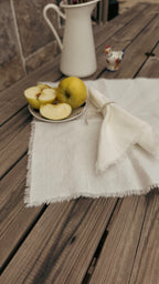 A rustic white linen tablecloth with frayed edges laid out on a weathered wooden table, styled with a small plate of sliced yellow apples, a folded napkin, a white ceramic pitcher, and a small decorative chicken figurine.