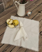 A white fringed linen napkin tied with a silver ring, placed on a rustic wooden table alongside a small plate of sliced apples and a white pitcher filled with pink flowers.
