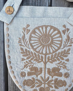 A close-up of a light gray linen apron or fabric bag laid flat on a weathered gray wooden surface. The fabric features a prominent tan embroidered floral and bird motif, with a matching gray fabric strap and coconut shell button at the top left.