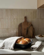 A kitchen countertop scene with two golden-brown bread rolls in a black baking dish, draped with a light beige linen cloth. A second folded linen cloth sits beside the dish, and two wooden cutting boards lean against a beige tiled backsplash.