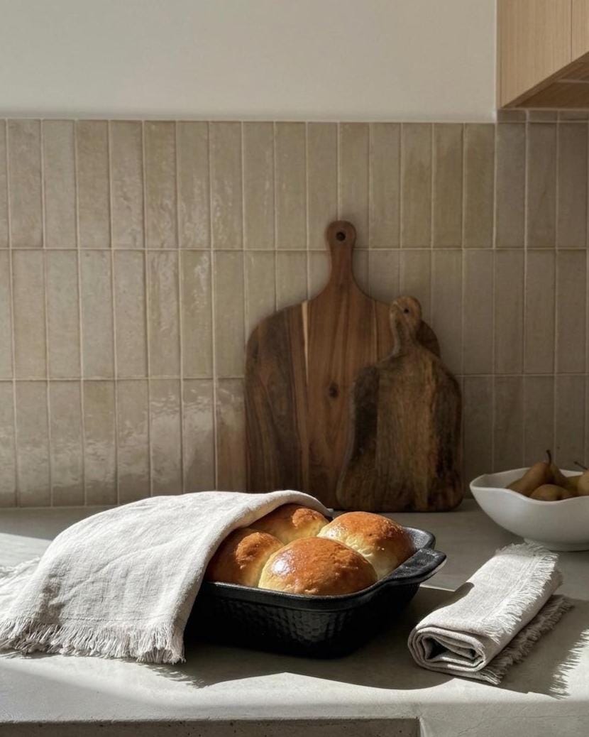 A kitchen countertop scene with two golden-brown bread rolls in a black baking dish, draped with a light beige linen cloth. A second folded linen cloth sits beside the dish, and two wooden cutting boards lean against a beige tiled backsplash.