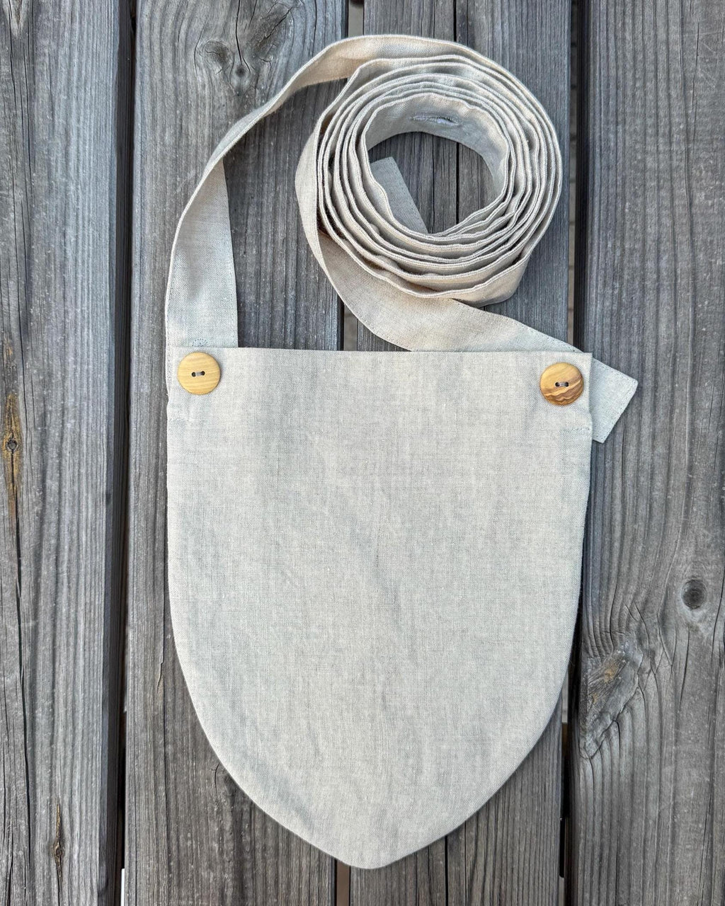 A light beige linen shoulder bag with a wide strap and  two coconut shell buttons, laid flat on a gray wooden plank background.