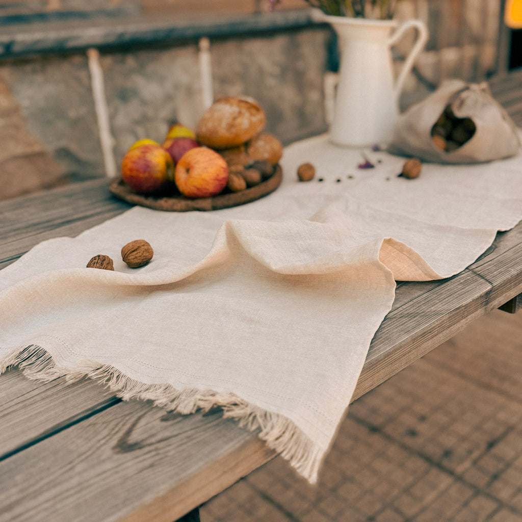 A rustic outdoor table styled with a cream-colored fringed tablecloth, a white pitcher of purple flowers, a small wooden board with bread and fruit, and scattered walnuts, set against a warm beige wall.