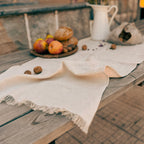 A rustic outdoor table styled with a cream-colored fringed tablecloth, a white pitcher of purple flowers, a small wooden board with bread and fruit, and scattered walnuts, set against a warm beige wall.