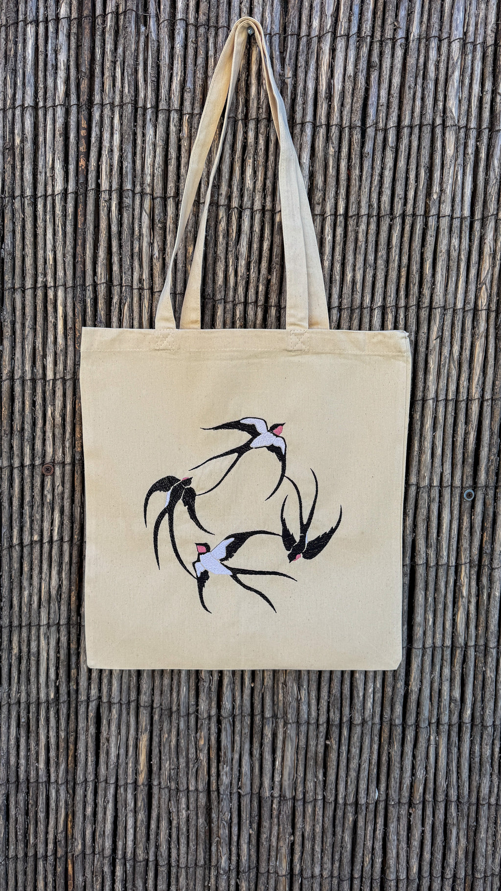 A natural beige canvas tote bag with embroidered swallow birds, hanging against a dark brown bamboo wall.