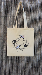 A natural beige canvas tote bag with embroidered swallow birds, hanging against a dark brown bamboo wall.