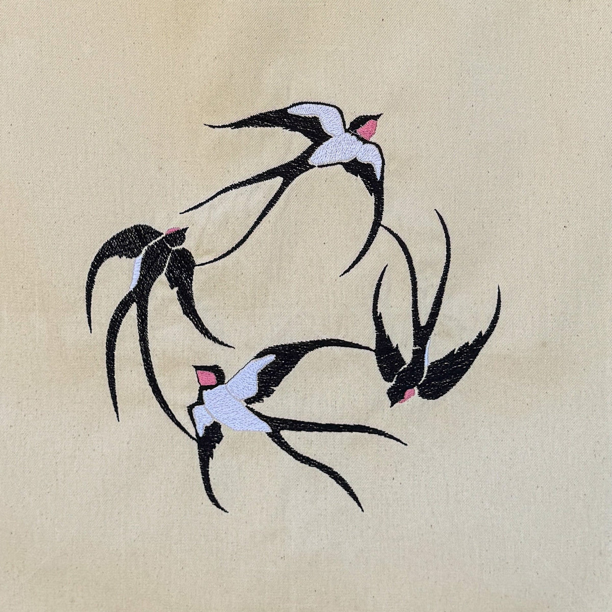 A natural beige canvas tote bag with embroidered swallow birds, hanging against a dark brown bamboo wall.