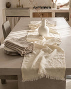 A neutral-toned dining table styled with a cream linen table runner, matching folded napkins, white plates, and a white ceramic pitcher in a softly lit modern kitchen.