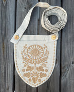 A light beige linen shoulder bag with a shield-shaped front pocket featuring a tan floral embroidery and button closures, shown on a dark gray wooden plank background.