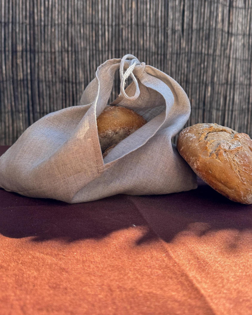 A natural linen drawstring bread bag containing a loaf of bread, with another loaf resting beside it on a dark brown surface against a woven reed background.