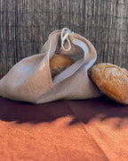 A natural linen drawstring bread bag containing a loaf of bread, with another loaf resting beside it on a dark brown surface against a woven reed background.