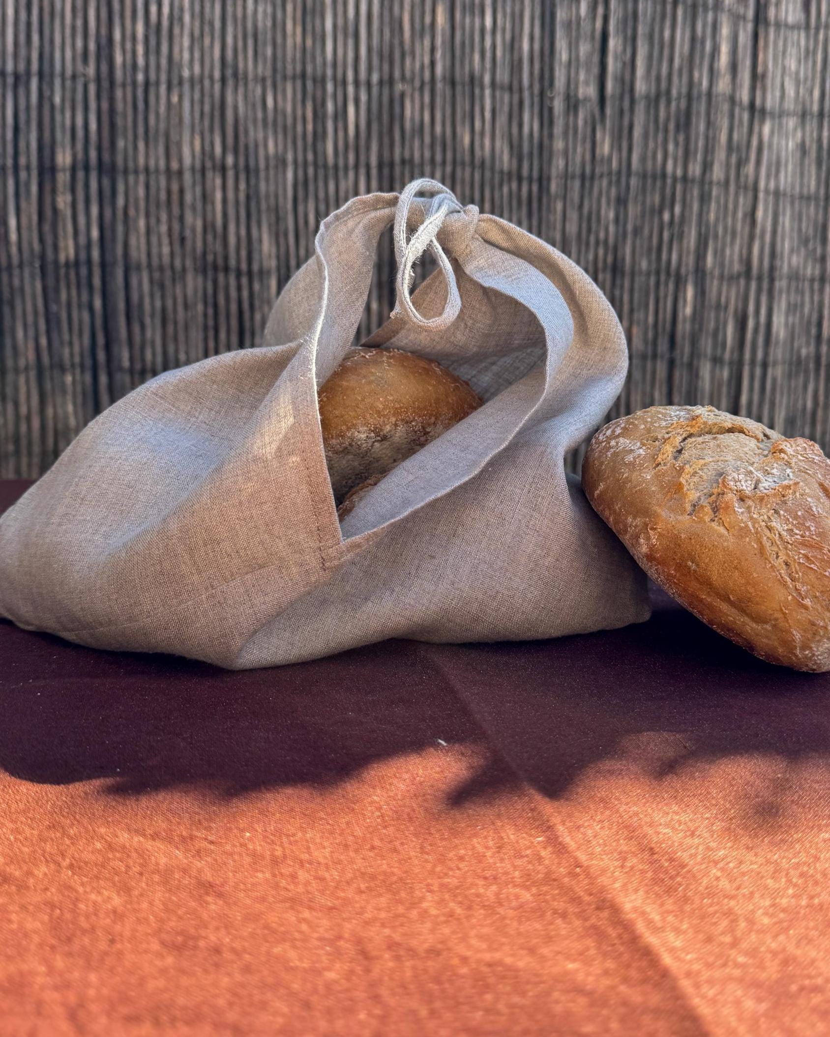 A natural linen drawstring bread bag containing a loaf of bread, with another loaf resting beside it on a dark brown surface against a woven reed background.