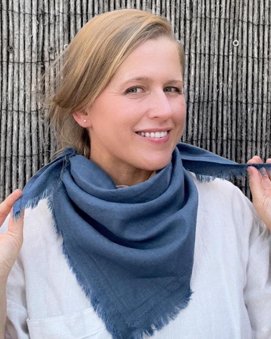 A woman wearing a navy blue square scarf with fringed edges, holding the corners to show its drape, set against a rustic bamboo wall.