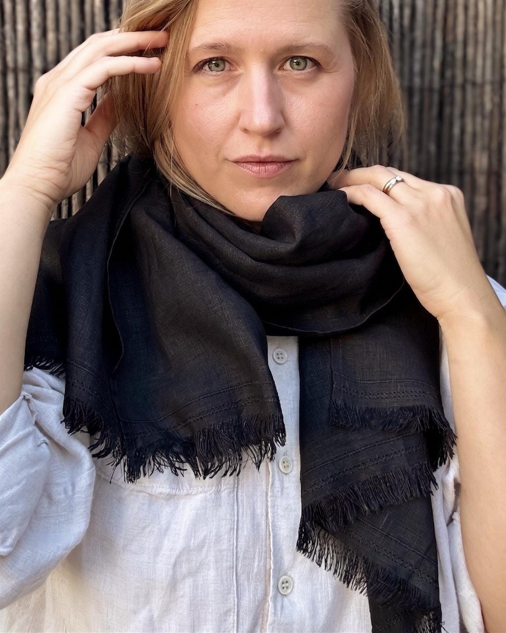 A woman wearing a black fringed scarf wrapped around her neck, paired with a white button-up shirt, set against a dark textured background.