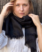 A woman wearing a black fringed scarf wrapped around her neck, paired with a white button-up shirt, set against a dark textured background.