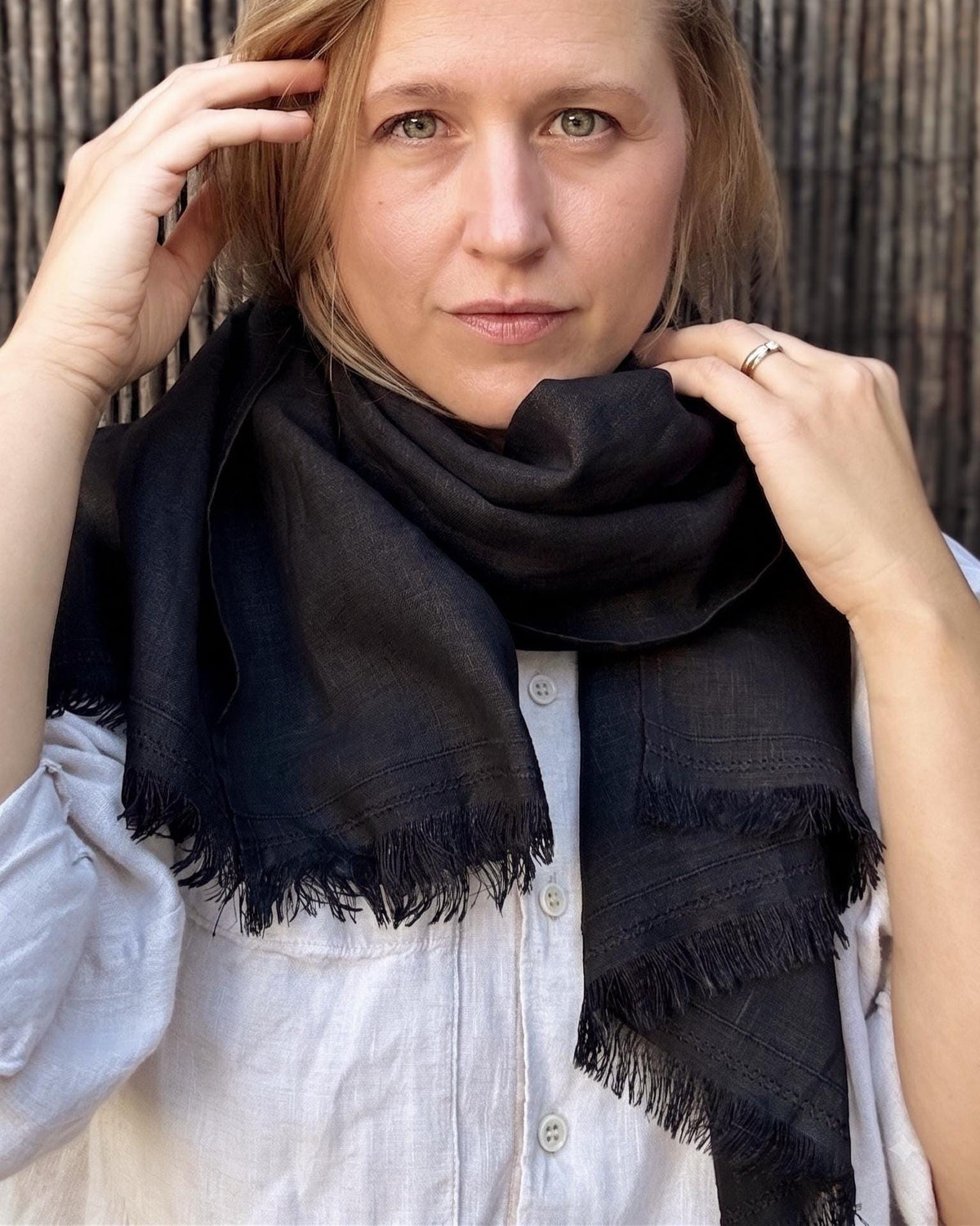 A woman wearing a black fringed scarf wrapped around her neck, paired with a white button-up shirt, set against a dark textured background.