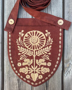 A rustic brown linen pouch with a shield-shaped body and a wide strap, decorated with cream-colored embroidery of a sunflower, birds, and floral motifs. The pouch is laid flat on a weathered gray wooden background.