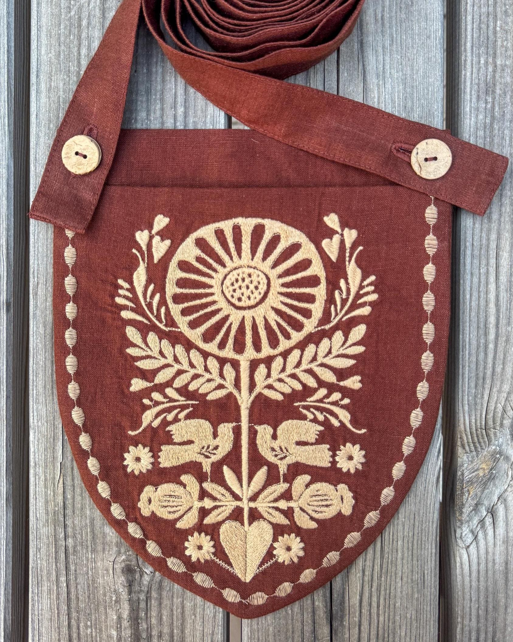A rustic brown linen pouch with a shield-shaped body and a wide strap, decorated with cream-colored embroidery of a sunflower, birds, and floral motifs. The pouch is laid flat on a weathered gray wooden background.