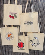 Five natural canvas tote bags with embroidered Ukrainian designs are displayed against a dark brown woven reed background. The designs include cherries, a fish, a bird, a strawberry, and a cursive name 'Mamina'.