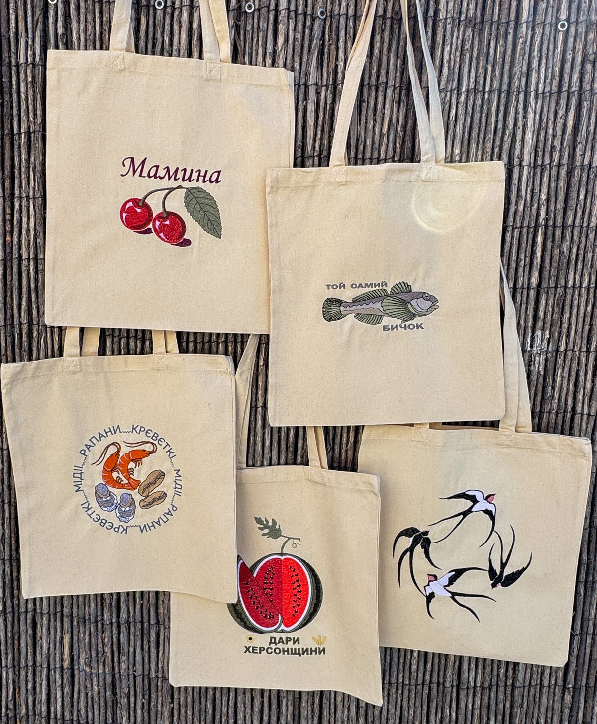 Five natural canvas tote bags with embroidered Ukrainian designs are displayed against a dark brown woven reed background. The designs include cherries, a fish, a bird, a strawberry, and a cursive name 'Mamina'.