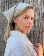 A woman wearing a light beige linen headscarf and a matching linen shirt, posing in front of a vertical wicker screen.
