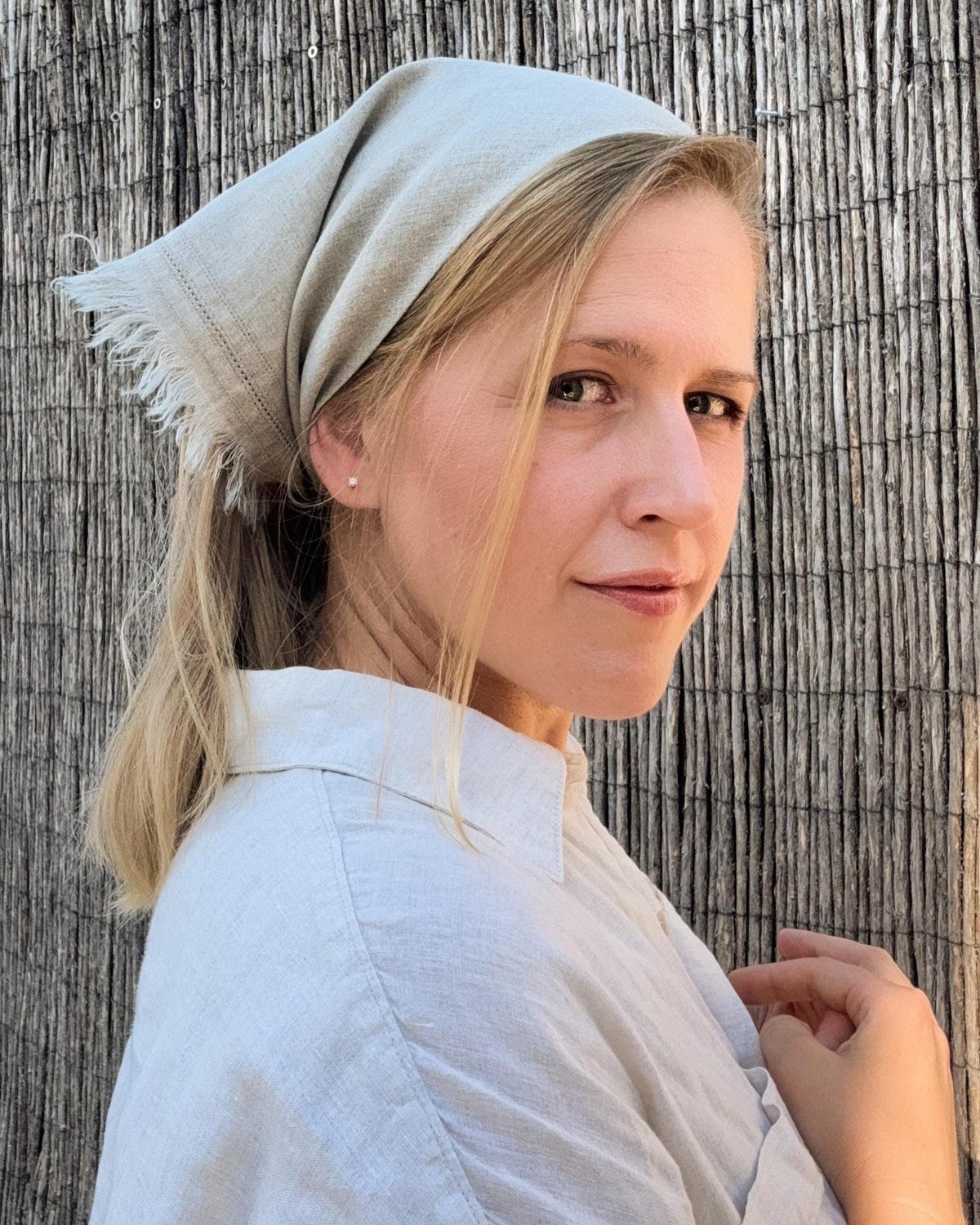 A woman wearing a light beige linen headscarf and a matching linen shirt, posing in front of a vertical wicker screen.