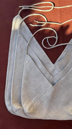 A close-up of a light gray linen-textured garment, likely a camisole or top, with layered V-neck panels and thin white drawstrings, laid flat on a dark reddish-brown background.