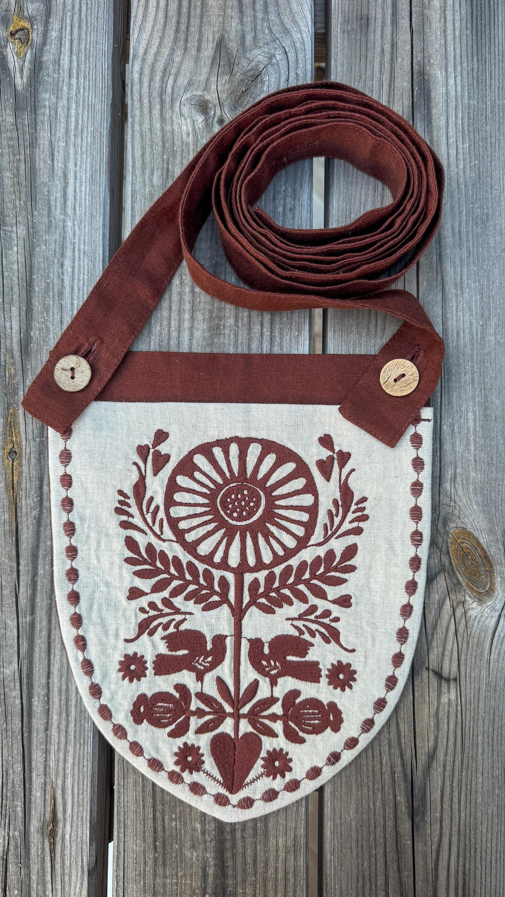 A rustic linen shoulder bag with a brown strap and a shield-shaped body, featuring a brown floral and bird embroidery on an off-white linen background, displayed on a weathered gray wooden surface.