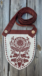 A rustic linen shoulder bag with a brown strap and a shield-shaped body, featuring a brown floral and bird embroidery on an off-white linen background, displayed on a weathered gray wooden surface.