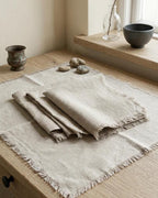 A set of natural beige linen table linens, including a flat rectangular placemat and folded napkins with fringed edges, styled on a light wood table next to a window with decorative stones and ceramic vessels.